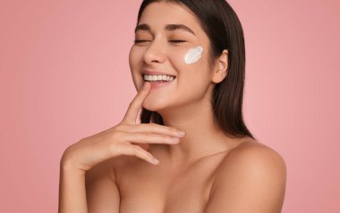 Beautiful young female with closed eyes smiling and touching chin while applying cream on face on pink background in studio