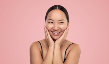 Positive young ethnic female smiling and touching face while looking at copy space isolated on pink background in studio