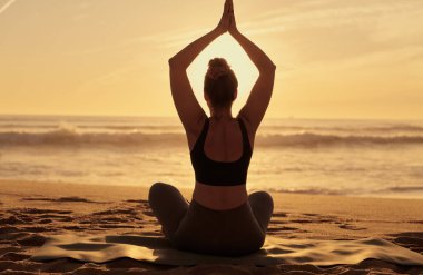 Back view silhouette of unrecognizable female in sportswear sitting on sandy beach and stretching arms, while practicing yoga in lotus pose and meditating near ocean