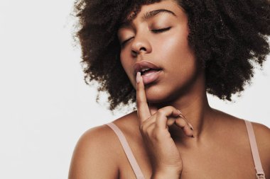 Sensual black female with Afro hairstyle touching lips and closing eyes during skin care routine against gray background