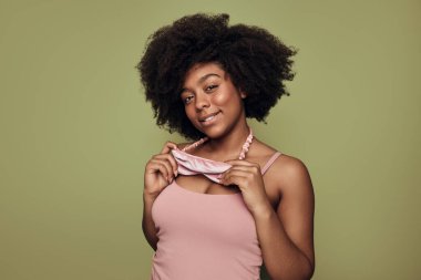 Optimistic young African American female with dark curly hair in pink top and sleep mask on neck, smiling and looking at camera against green background