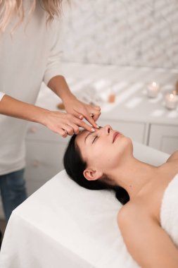Side view of crop anonymous female massaging forehead of young female client with closed eyes, while working in modern salon