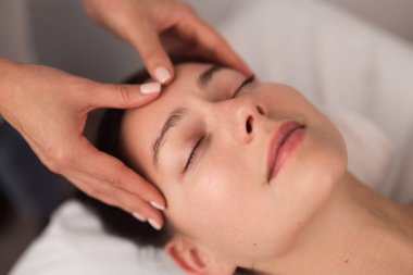 From above of crop unrecognizable cosmetician massaging forehead of young female client lying on couch with closed eyes during facial procedure in light salon