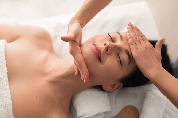Top view of crop unrecognizable masseuse massaging face of content young female customer with dark hair and bare shoulders, lying on table and smiling with closed eyes during beauty procedure