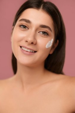 Positive young female with bare shoulders and cream smiling and looking at camera on pink background during skin care routine