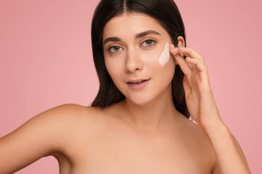 Beautiful young female with perfect skin applying moisturizing cream on face while looking at camera on pink background. Beauty and skincare concept