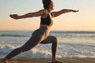 Side view of young concentrated barefooted female with dark hair in activewear performing Virabhadrasana B pose while practicing yoga near wavy ocean at sunset