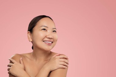 Optimistic Korean female with perfect skin looking away with smile and touching bare shoulders during spa session against pink background