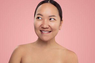 Curious young Asian female model with perfect skin and bare shoulders biting lip and looking away against pink background