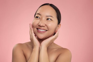 Delighted young Asian female model with dark hair and perfect skin, smiling and looking away while touching cheeks after skin care procedure against pink background
