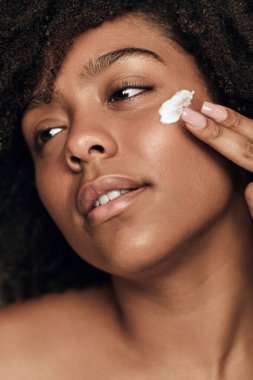 Young African American female with curly hair smearing moisturizing cream on cheek and looking away during skin care routine