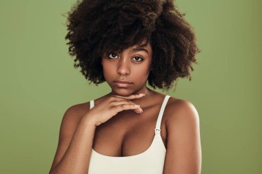 Self assured young African American female model with curly dark hair and perfect skin, in white bra touching chin and looking at camera against green background