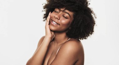 Side view of positive young African American female model with curly dark hair smiling with closed eyes while touching face after skincare procedure