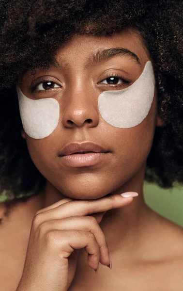 Crop young confident African American female model with dark curly hair perfect skin and eye patches, touching chin and looking at camera against green background