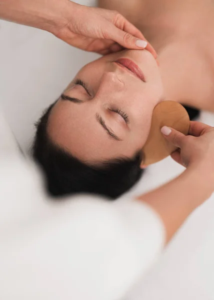 From above of crop anonymous cosmetologist massaging face of relaxed young female client with wooden gua sha tool, during skin care procedure in light salon
