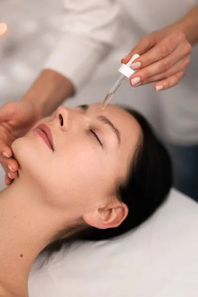 From above of crop anonymous cosmetician applying acid serum on face of calm young female client, with perfect skin lying on massage table with closed eyes during skincare procedure