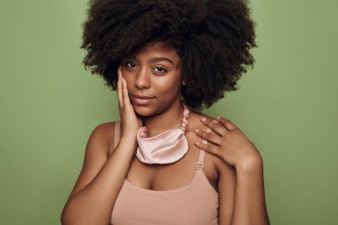 Afro saçlı, yumuşak tenli, yeşil arka planda uyandıktan sonra pembe ipek uyku maskesi takan çekici Afro-Amerikalı kadın.