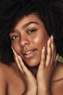 Kıvırcık saçlı, yüzüne dokunan ve siyah arka planda kameraya bakan genç bir Afro-Amerikan kadın.