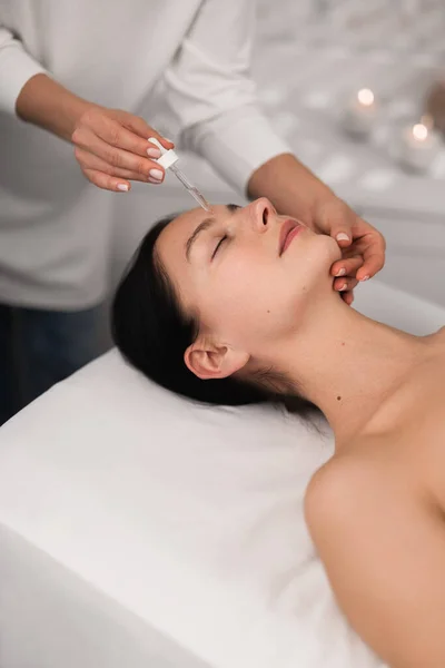 High angle of crop anonymous beautician applying serum with dropper on face of calm young female client, lying on table with closed eyes during skincare procedure in salon