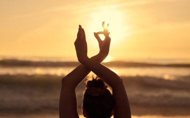 Siyah saçlı, güçlü dalgalı okyanusun önünde Gyan Mudra elleriyle dikilip günbatımında gökyüzüne karşı yoga yapan tanınmamış bir kadının arka görüntüsü.