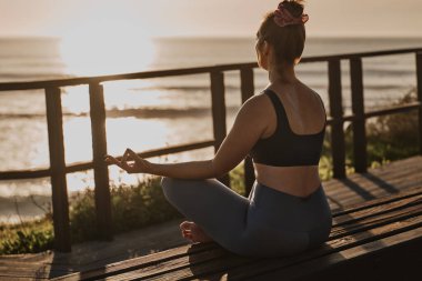Nilüfer ağacından bir bankta oturup, gün batımına karşı dalgalı okyanusun yanında meditasyon yapan, Lotus 'ta çıplak ayaklı bayanın tüm vücudu.