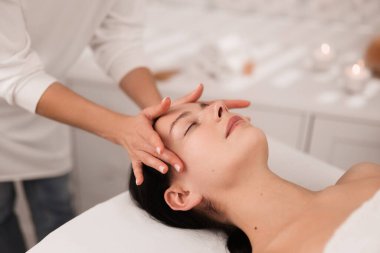 Side view of young female client lying on table with closed eyes in beauty salon while masseur doing head massage