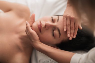 From above crop masseuse massaging face of calm female client with closed eyes during work in spa salon
