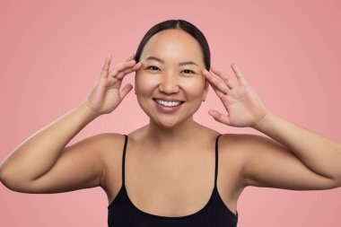 Young Asian lady in black top touching soft and fresh skin of face and looking at camera with smile on pink background