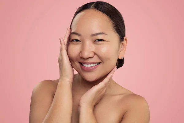 幸せな韓国の女性で裸の肩は滑らかな顔の肌に触れ ピンクの背景に笑顔でカメラを見て — ストック写真