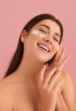 Delighted young female millennial with dark hair and bare shoulders, smiling happily with closed eyes while applying moisturizing cream on face during during skin care routine against pink background