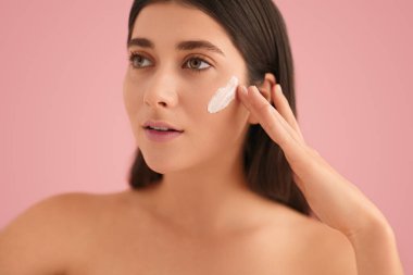 Young brunette with bare shoulders smearing cream on cheek and looking away during beauty routine against pink background