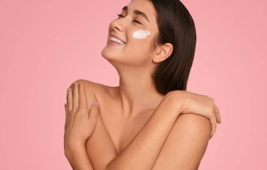 Happy young woman with cream on cheek smiling with closed eyes and touching bare shoulders during skin care routine against pink background