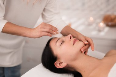 Crop anonymous masseur doing facial massage to young woman client lying on table with closed eyes. Specialist using gua sha professional tool for therapy procedure