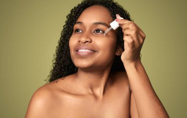 Positive half naked African American female with curly hair and bare shoulders smiling looking up while applying serum under eye against green background