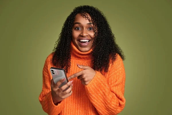 Excited young black female in knitted sweater smiling and looking at camera while pointing at cellphone screen against green background