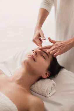 Anonim kozmetik uzmanı güzellik tedavisi sırasında gözleri kapalı bir şekilde masaya uzanan genç bayan müşteriyi sakinleştirmek için akupunktur yüz masajı yapıyor.