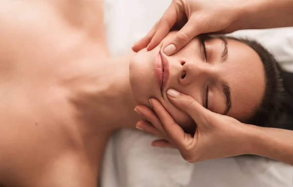 Top view of crop anonymous professional massage therapist massaging face of relaxed female client with closed eyes during procedure in spa salon