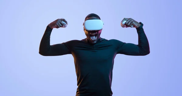 興奮した若いアフリカ系アメリカ人男性ゲーマーでVrメガネゲームパッドの叫びと青い背景に対する勝利を祝いながら 双眼鏡を示します — ストック写真