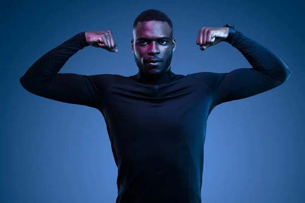 自信自信的年轻的非洲裔美国男性运动员 身穿时髦的黑色运动服 展示二头肌 并在蓝色背景下看着相机 — 图库照片