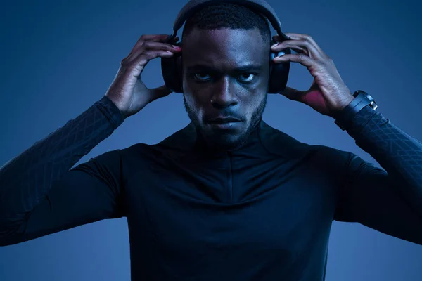 有自信的年轻非洲裔美国男性运动员 穿着深色的运动服 调整耳机 一边听音乐一边在蓝色工作室听音乐 — 图库照片