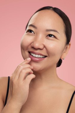 Pembe arka plana bakarken parmaklarıyla dudaklarına ruj süren genç, pozitif etnik bir kadın.