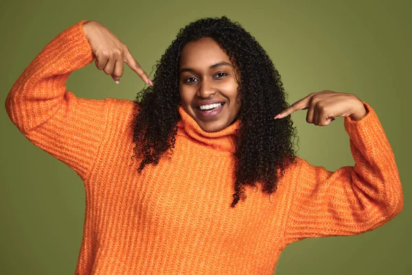 身穿橙色毛衣 卷发的非洲裔美国女性 面带微笑 用手指指着自己的绿色背景 — 图库照片