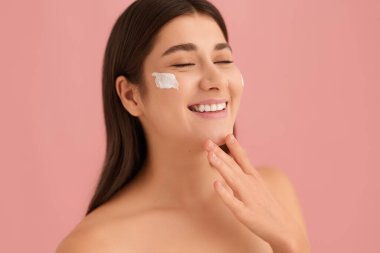 İyimser genç kadın, yanağında nemlendirici krem, çenesine dokunuyor ve pembe arka planda güzellik sırasında kapalı gözlerle gülümsüyor.