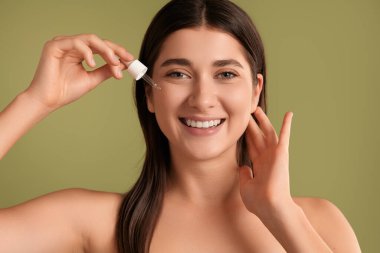 Çıplak omuzlu neşeli genç esmer. Yüzüne pipetle nemlendirici sıvı döküyor ve yeşil arka plandaki kameraya bakıyor.