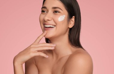 Çıplak omuzlu, hassas bir kadın. Yanağına krem sürmüş. Çenesine dokunmuş, pembe arka planına bakmıyor.