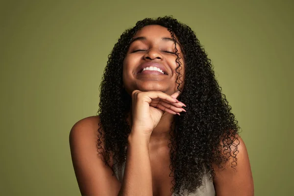 Young African American woman with curly hair leaning chin on hand and closing eyes while laughing on green background