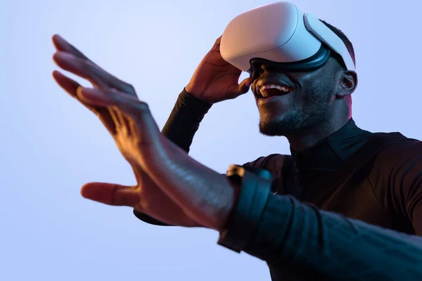 Delighted African American bearded man in black turtleneck and white virtual reality headset interacting with cyberspace while standing on blue background