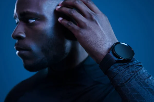 Crop self assured young African American male millennial in black activewear and smart watch adjusting wireless headphones and looking away against blue background