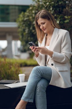 Takım elbiseli kadın girişimci netbook ile gülümsüyor ve şehir caddesinde uzaktan çalışırken cep telefonunun ekranına bakıyor.