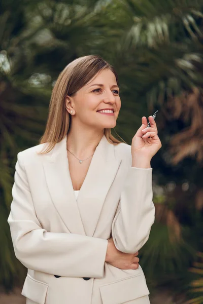 Confident positive elegant young female writer in beige suit with long blond hair smiling and looking away, while standing in tropical park with pen in hand